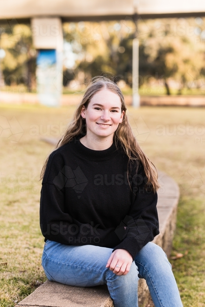 Young woman sitting on brick step in park smiling - Australian Stock Image