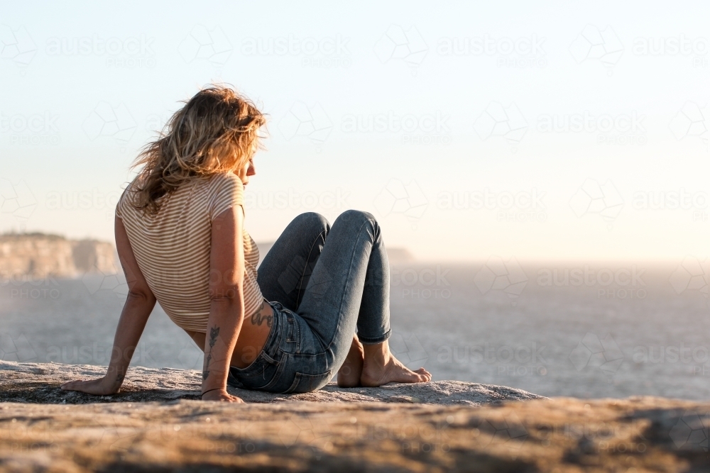 Young woman sitting on coastal clifftop at sunrise looking out to sea - Australian Stock Image