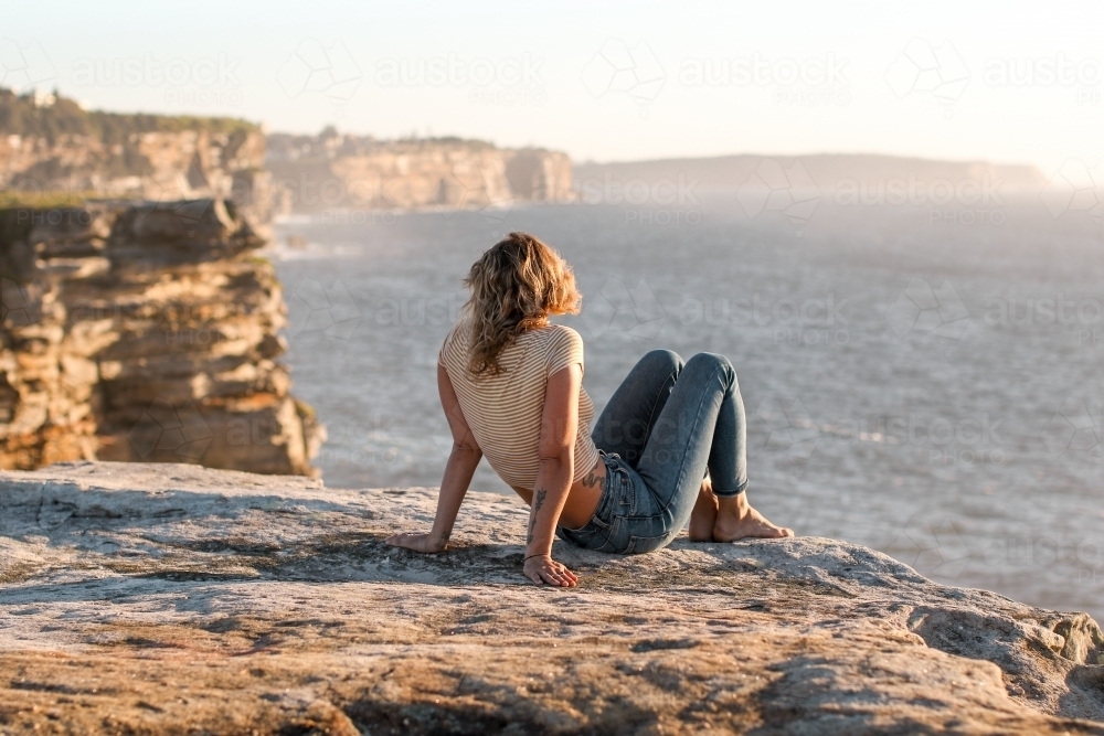 Young woman sitting on coastal clifftop at sunrise looking out to sea - Australian Stock Image