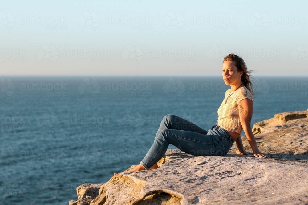 Young woman sitting on coastal clifftop at sunrise looking out to sea - Australian Stock Image