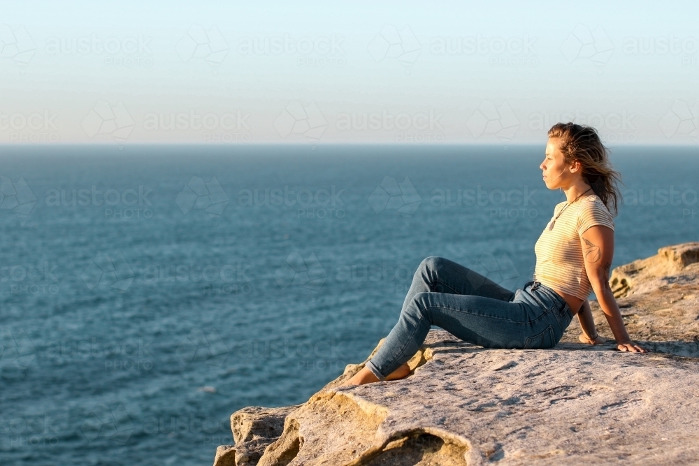 Young woman sitting on coastal clifftop at sunrise looking out to sea - Australian Stock Image