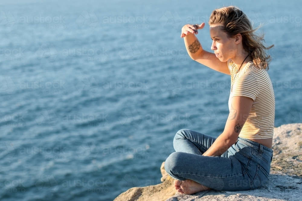 Young woman sitting on coastal clifftop at sunrise looking out to sea - Australian Stock Image