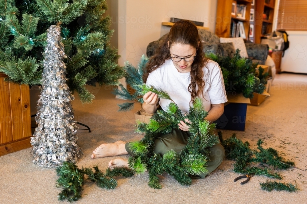 Young woman sitting on floor making Christmas wreath from branches of old Christmas tree - Australian Stock Image