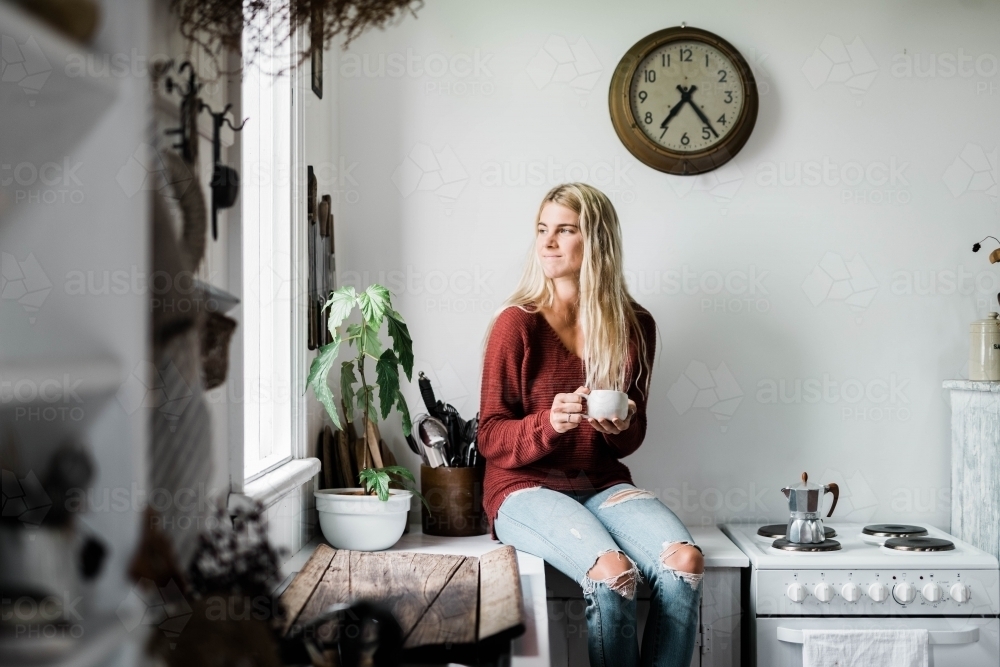 Young woman sitting on kitchen bench drinking coffee - Australian Stock Image