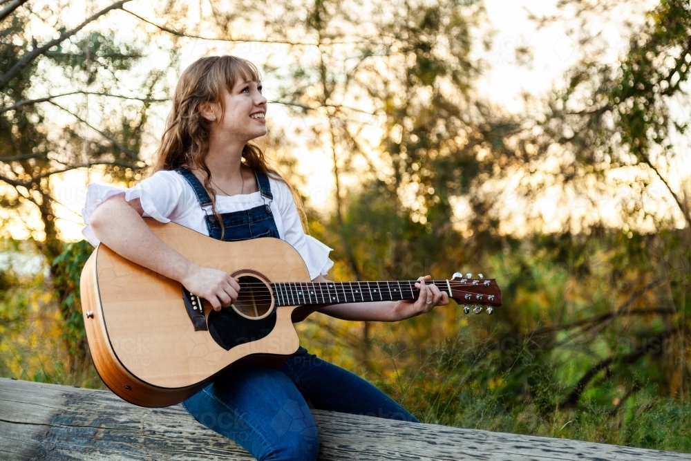 Young woman sitting on log playing instrument - Australian Stock Image