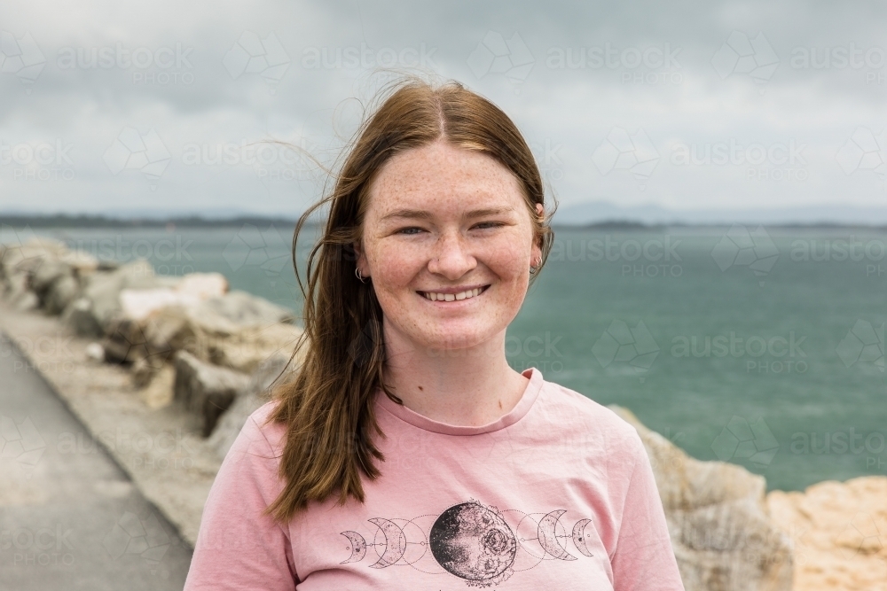 Young woman smiling standing on rock wall with ocean water behind - Australian Stock Image
