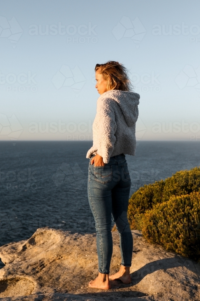 Young woman standing on coastal clifftop at sunrise looking out to sea - Australian Stock Image