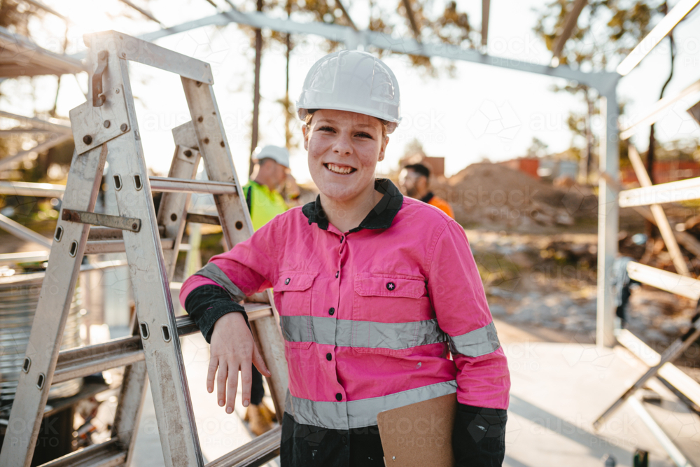 Young woman standing on constructions site while leaning on a steel ladder. - Australian Stock Image