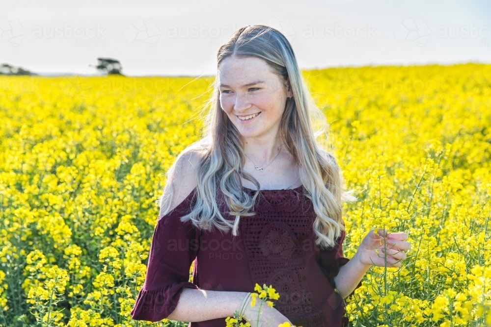 Young woman standing smiling in yellow canola field in paddock on farm - Australian Stock Image