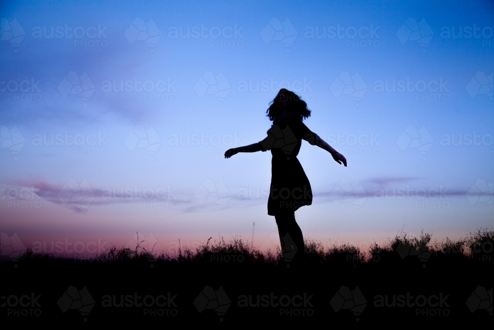 Young woman twirling silhouetted against pastel dusk sky - Australian Stock Image
