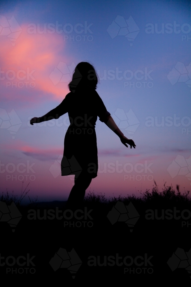 Young woman twirling silhouetted against pastel dusk sky - Australian Stock Image