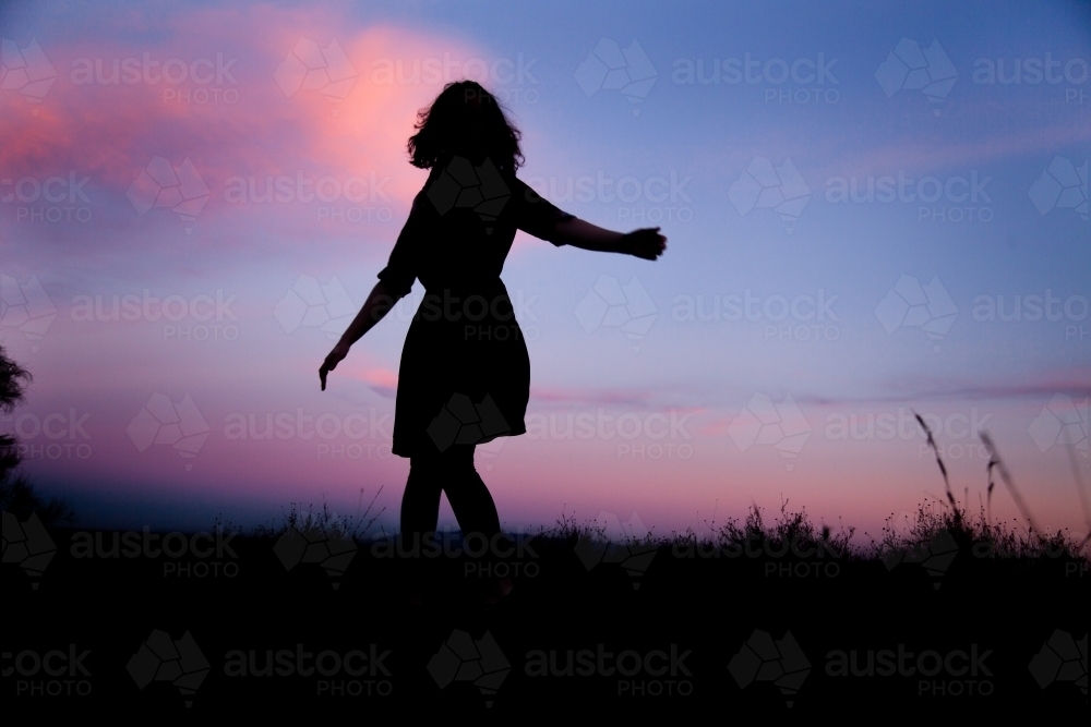 Young woman twirling silhouetted against pastel dusk sky - Australian Stock Image