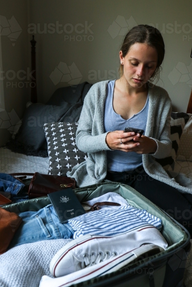 Young woman using her phone next to her packed bag ready to travel - Australian Stock Image