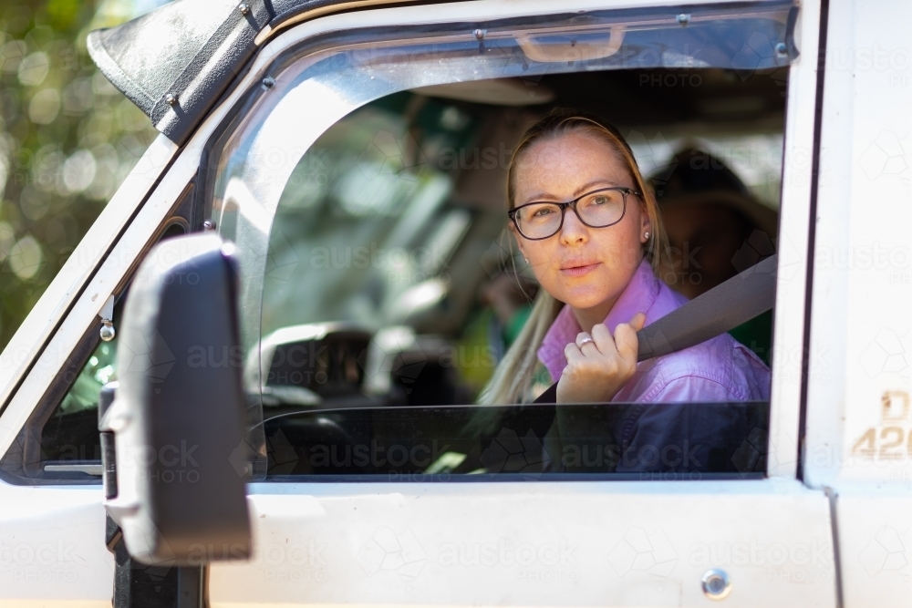 young woman wearing spectacles looking out car door while pulling on seatbelt - Australian Stock Image