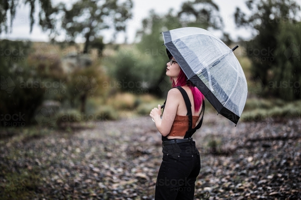 Young woman with bright pink hair looking up at rain holding umbrella - Australian Stock Image