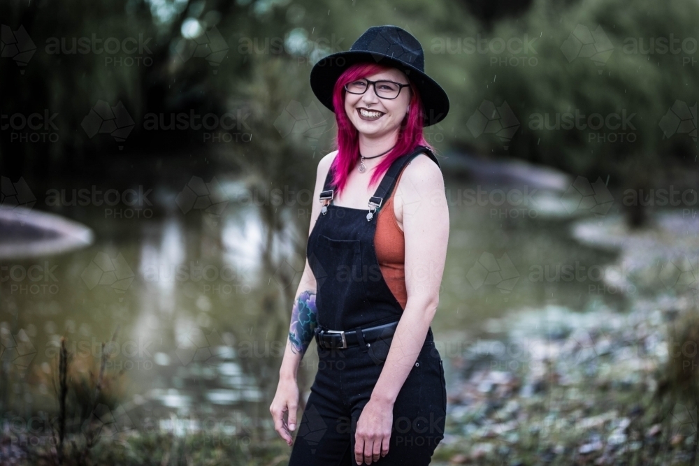 Young woman with coloured hair hat and glasses laughing near water - Australian Stock Image