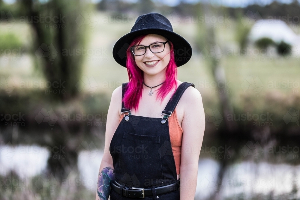 Young woman with pink hair glasses and hat smiling standing near river water - Australian Stock Image