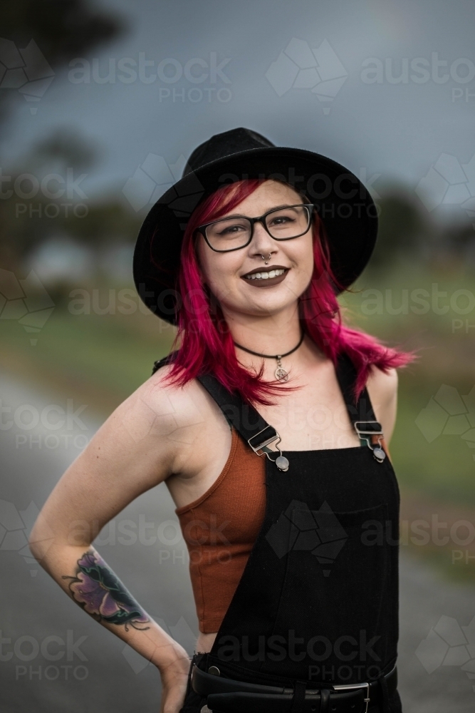 Young woman with pink hair hat glasses and tattoo smiling - Australian Stock Image