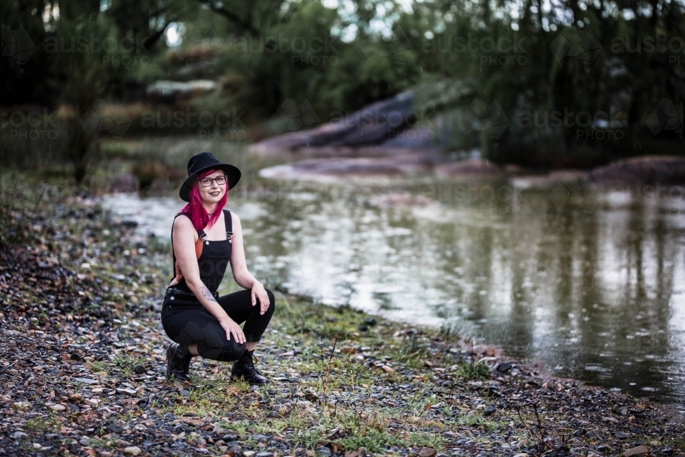 Young woman with pink hair kneeling on rocks near river water - Australian Stock Image