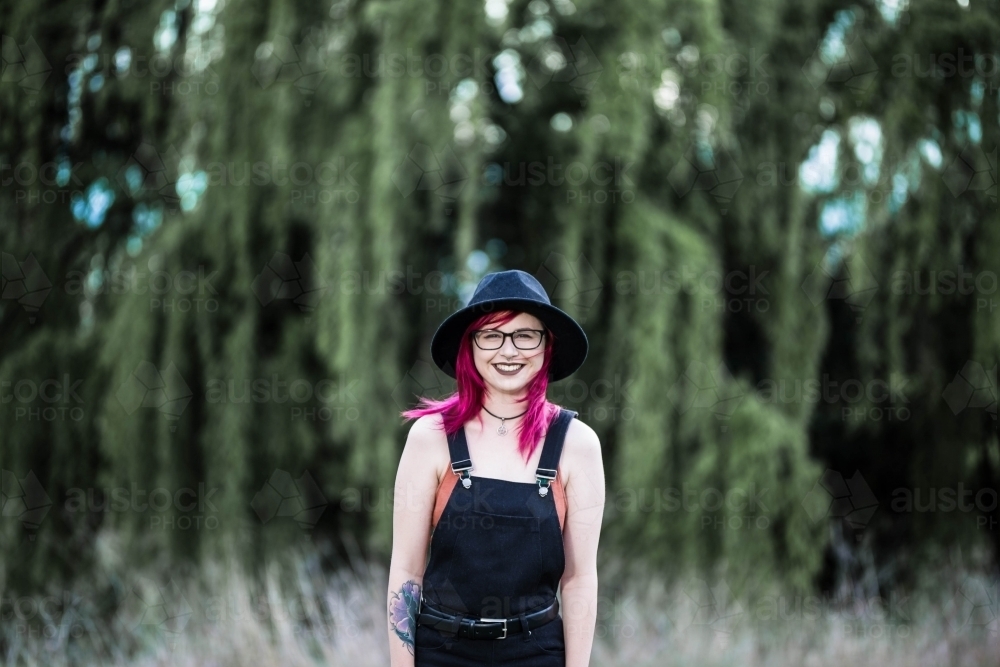 Young woman with pink hair smiling in front of willow tree - Australian Stock Image
