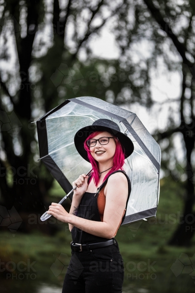 Young woman with pink hair standing in rain holding umbrella smiling - Australian Stock Image