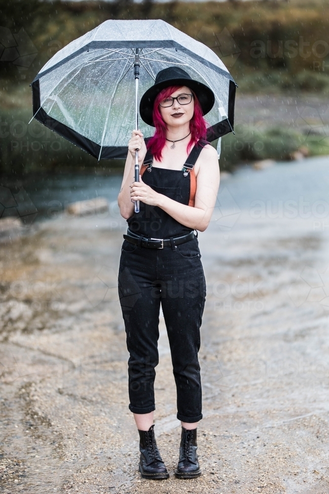 Young woman with pink hair standing near water holding umbrella in rain - Australian Stock Image