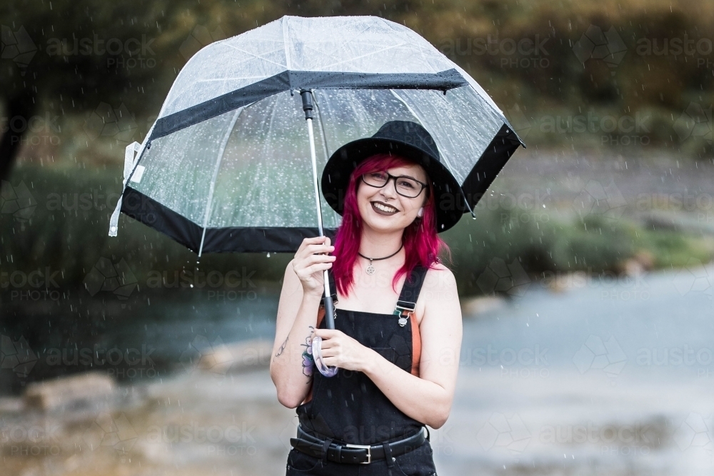 Young woman with pink hair standing near water in rain holding umbrella smiling - Australian Stock Image