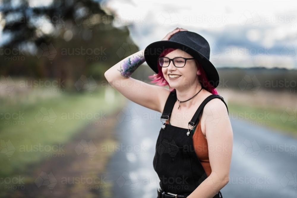 Young woman with pink hair tattoo and glasses holding onto hat as wind blows laughing happy - Australian Stock Image