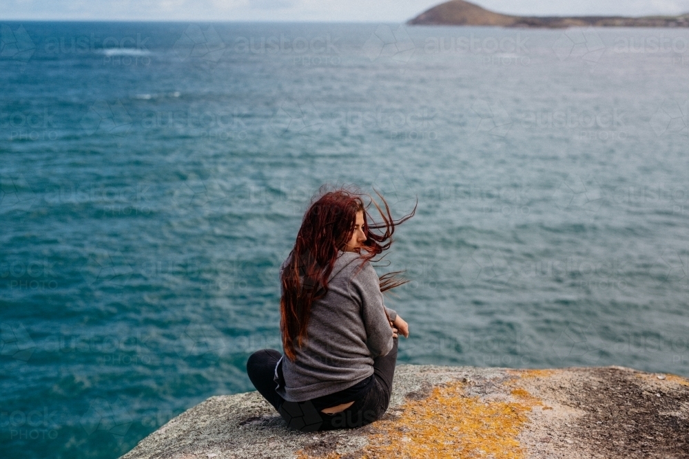 Young woman with red hair sitting on rock looking at ocean - Australian Stock Image