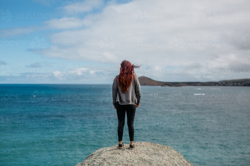 Young woman with red hair standing on rock looking at ocean - Australian Stock Image