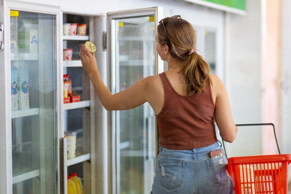 young woman with shopping basket taking yoghurt from shop refrigerator - Australian Stock Image