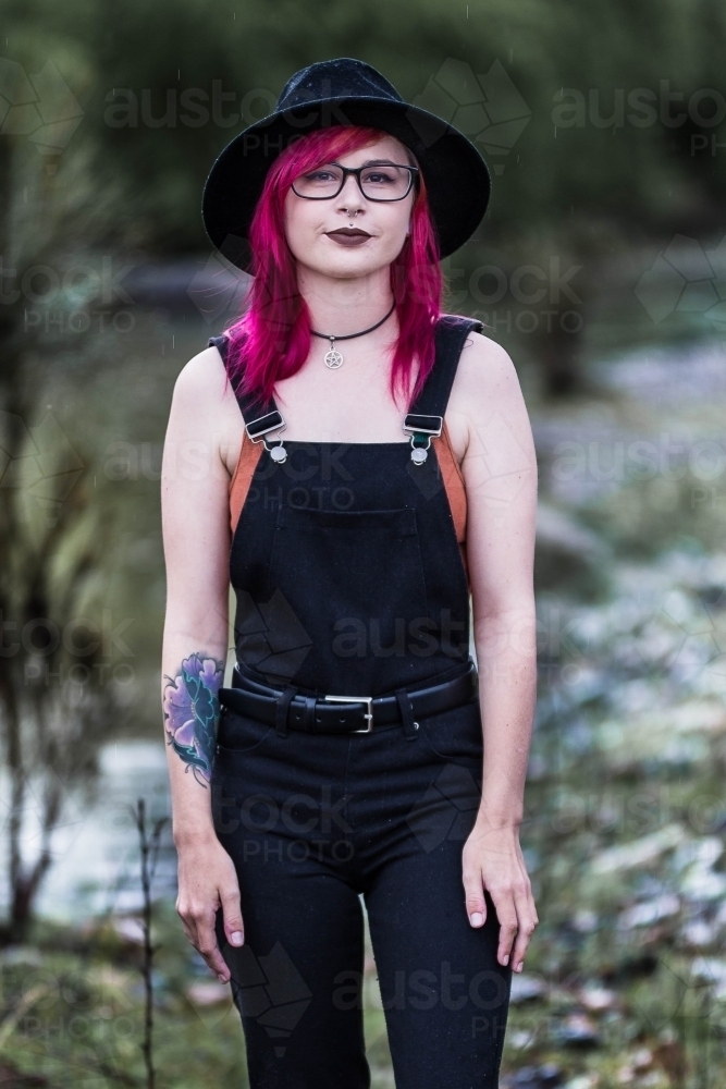 Young woman with tattoo pink hair hat and glasses standing near river - Australian Stock Image