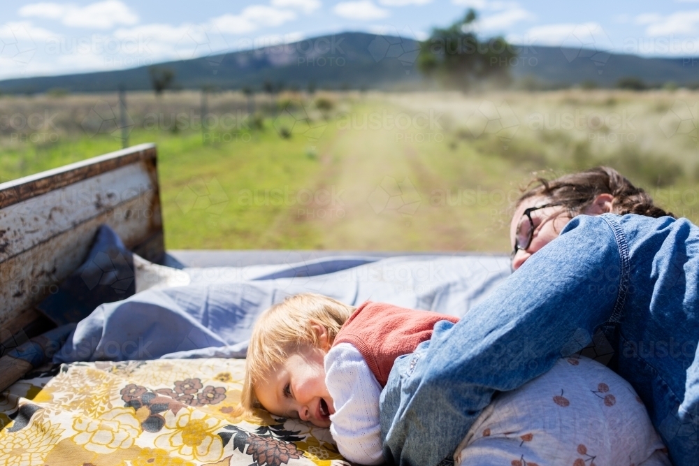 Young woman with toddler lying on cushions in back of ute traveling through paddock on country farm - Australian Stock Image