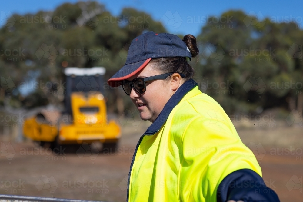 young woman working outdoors with hi-vis jacket and heavy machinery - Australian Stock Image