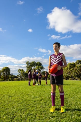 12 year old boy standing holding aussie rules football, with teammates in background - Australian Stock Image