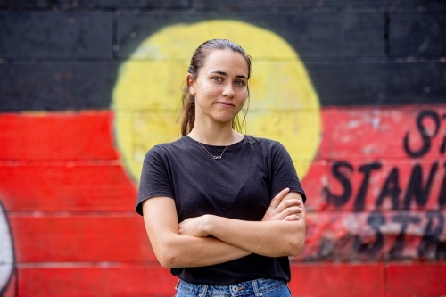 20-something Aboriginal woman standing with her arms crossed in front of a painted flag