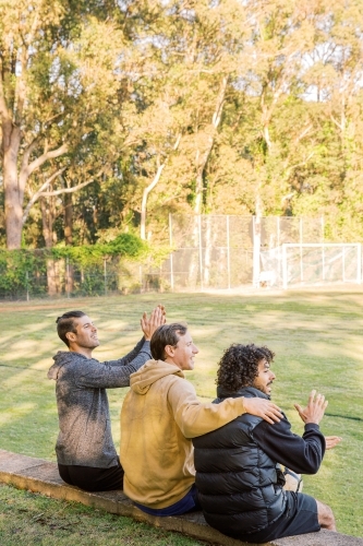 3 interracial young men sitting on a bench on the field looking a far smiling and cheering - Australian Stock Image