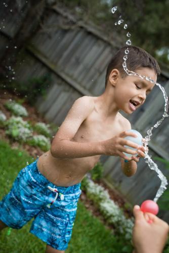 3 year old and 6 year old mixed race brothers play excitedly with water bombs in suburban backyard