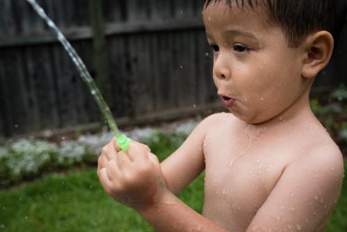 3 year old mixed race boy plays excitedly with water bombs in suburban backyard - Australian Stock Image