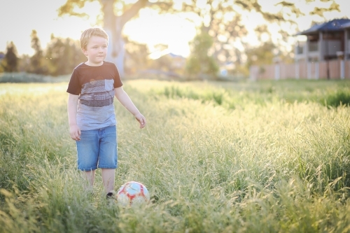 6 year old boy playing soccer in long grass at the park. Outdoor fun in nature, - Australian Stock Image
