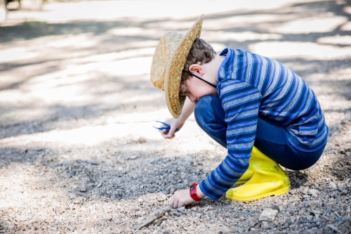 Child in gumboots and hat holding tweezers looking through rocks and dirt on ground