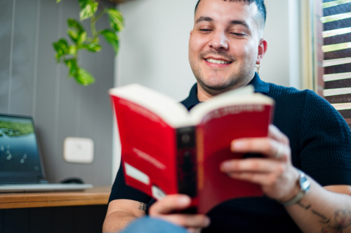 A australian man in his thirties is reading a book in a modern home office, smiling - Australian Stock Image