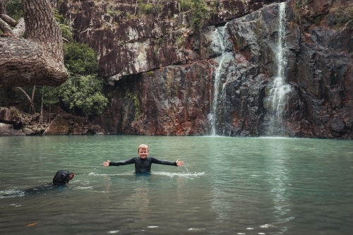 A boy and his dog swimming in a waterhole with trickling waterfall - Australian Stock Image