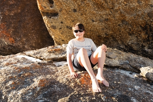 a boy child wearing sun glasses sitting on boulder rocks at Squeaky beach in Wilson's Promontory Sou - Australian Stock Image