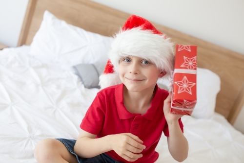 A boy wearing a red shirt sitting on a bed wearing a Santa hat shaking a wrapped christmas present, - Australian Stock Image