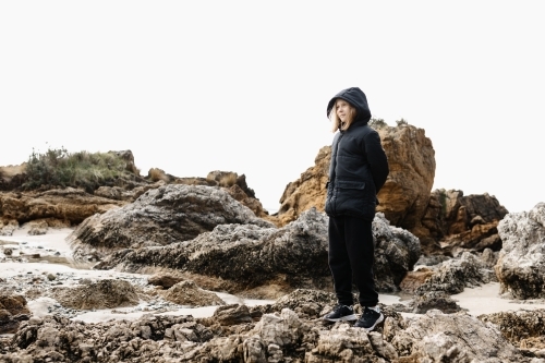A causcasian blonde girl wearing a warm jacket standing on rocks at a beach in winter - Australian Stock Image