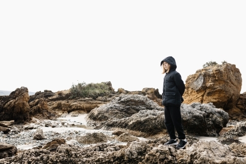 A causcasian blonde girl wearing a warm jacket standing on rocks at a beach in winter - Australian Stock Image