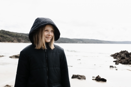 A causcasian blonde girl wearing a warm jacket walking on a beach in winter - Australian Stock Image