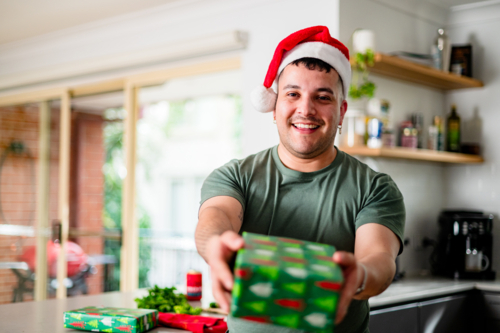 A cheerful man in a Santa hat shares a festive gift during the holiday season in his kitchen - Australian Stock Image