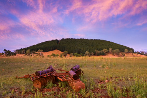 A colourful sunset sky over a rusty plough in a paddock in front of a tree covered mountain - Australian Stock Image